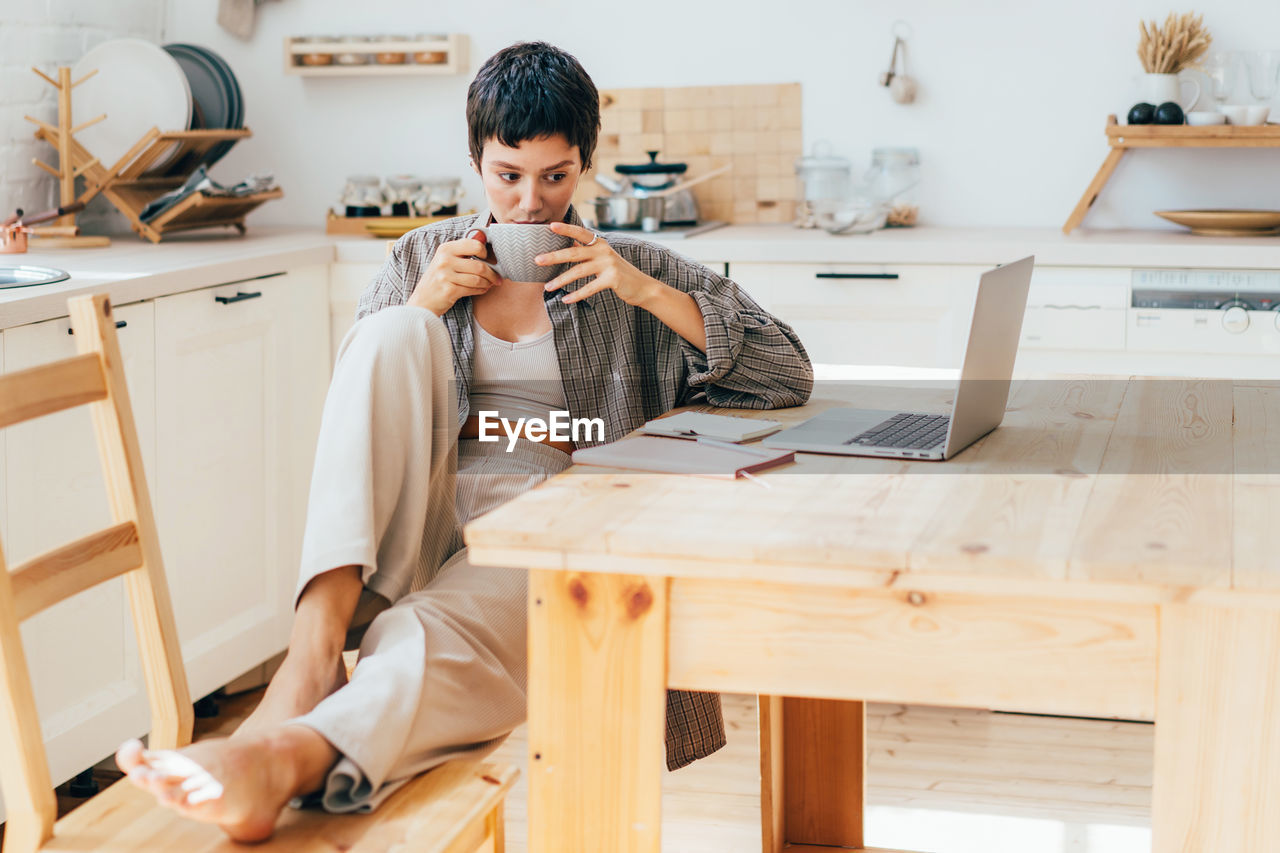 Young woman drinking coffee has a break while working on computer gadgets.