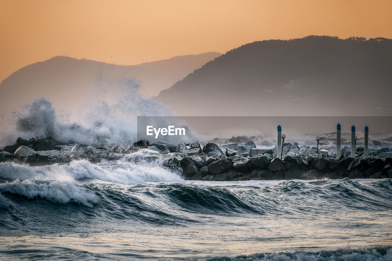 Waves splashing in sea against sky during sunset