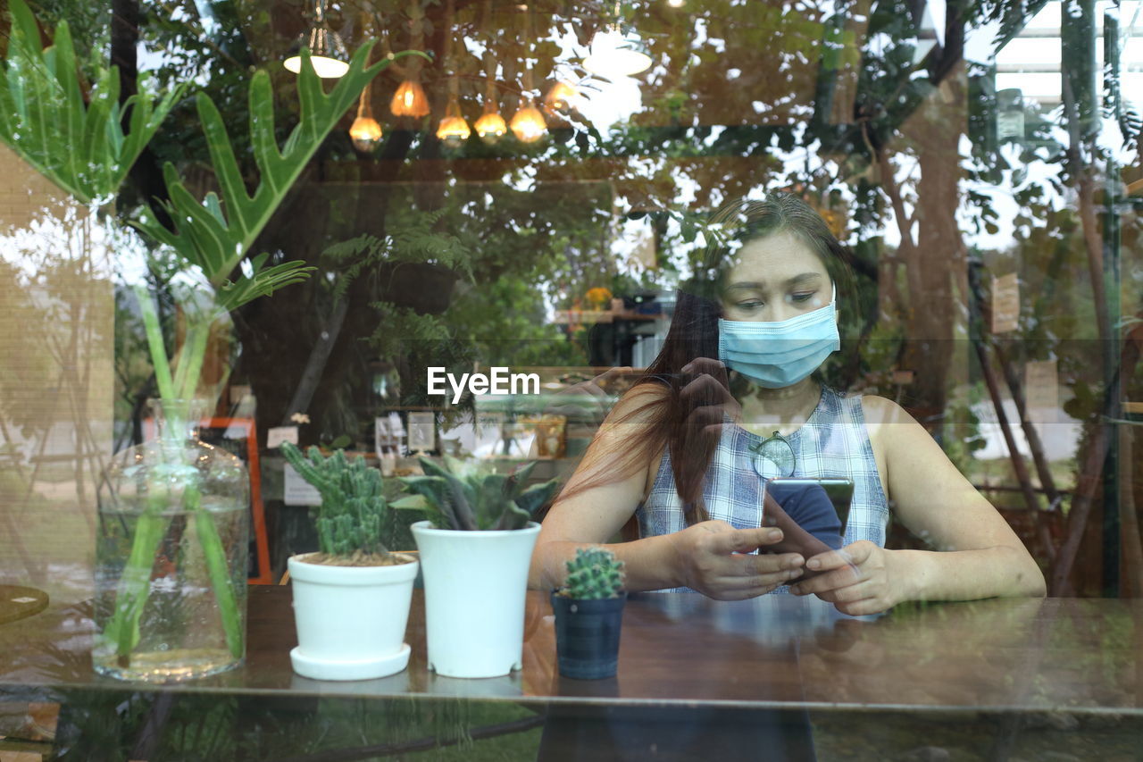 Woman wearing mask using mobile phone in cafe seen through window