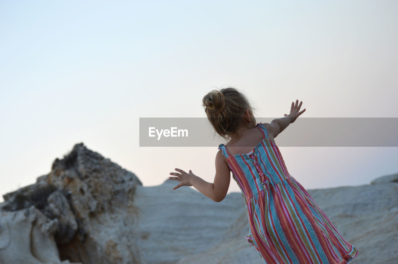 Rear view of girl standing by rock formation against clear sky during sunset