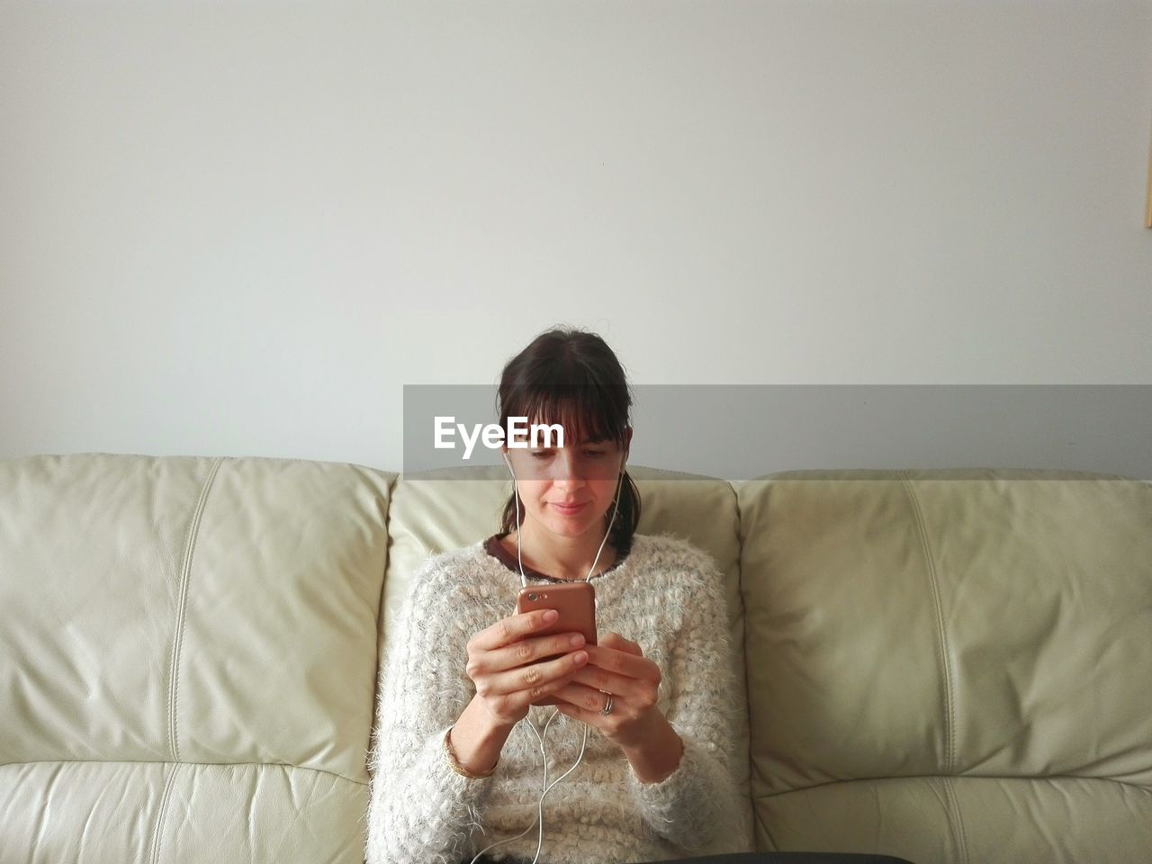 Woman listening music through smart phone while sitting on sofa against wall at home