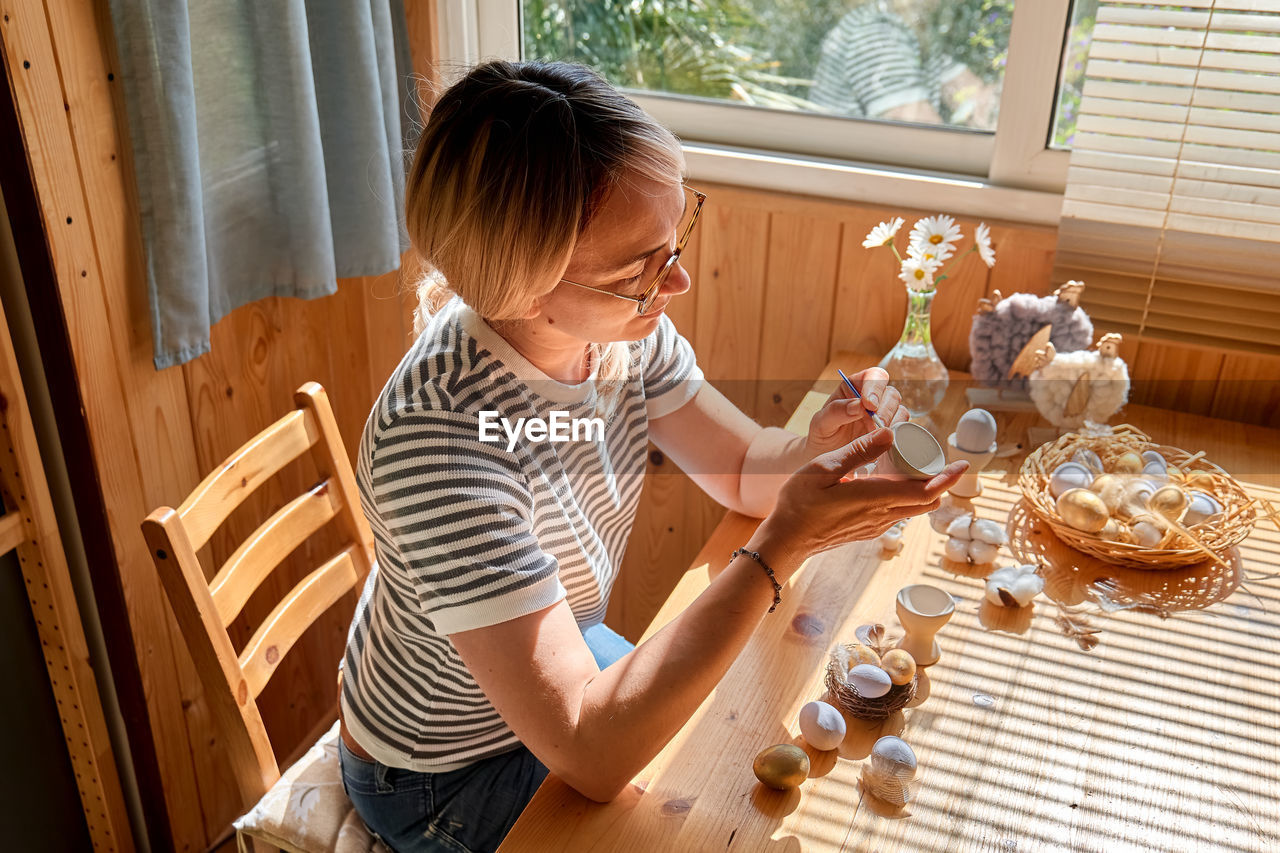 Blond woman preparing easter decoration at home, painting colorful easter eggs and coloring egg cups