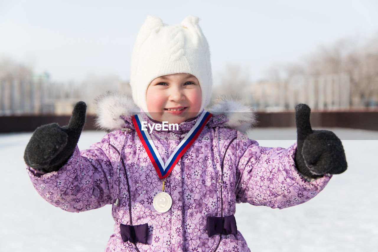 Portrait of smiling girl showing thumbs up while wearing medal standing against snow during winter