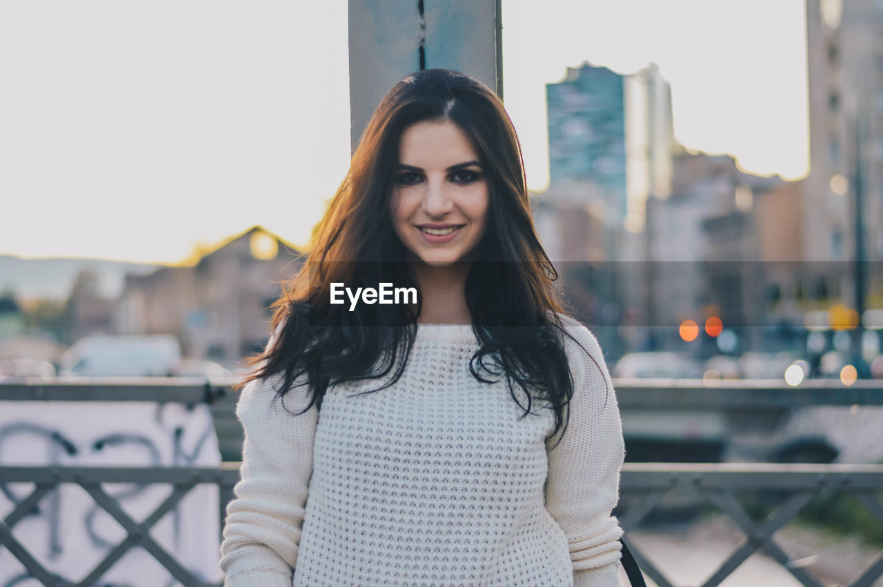 Portrait of smiling young woman standing against sky in city