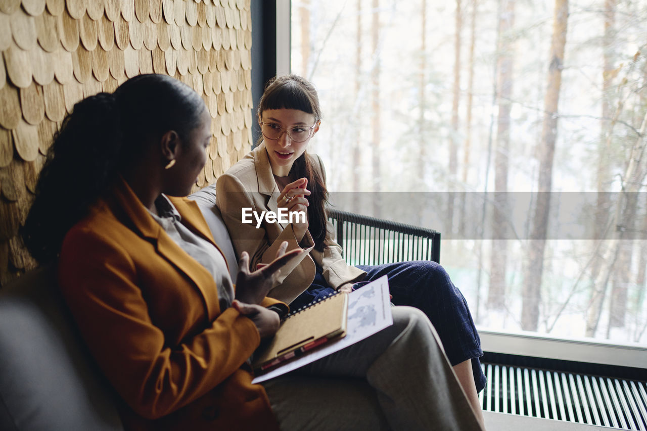 Businesswoman discussing with coworker sitting by glass window at convention center