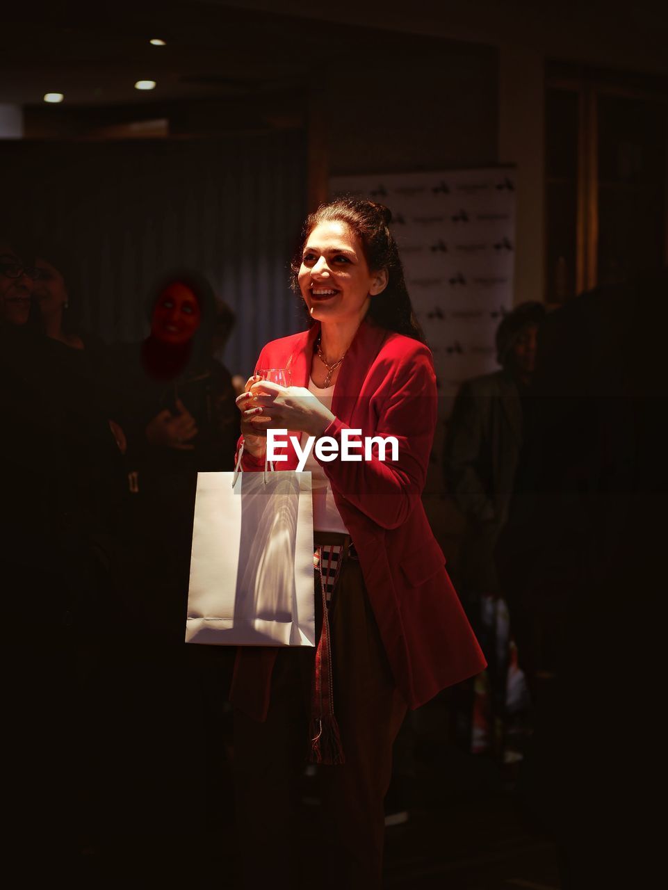 YOUNG WOMAN HOLDING CAMERA WHILE STANDING AGAINST GLASS