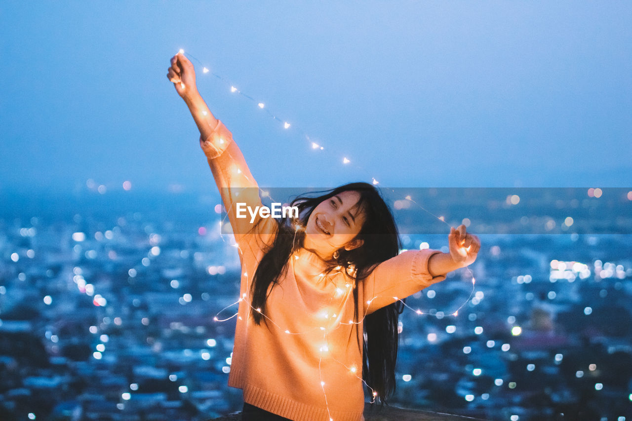 Cheerful woman holding illuminated string light against clear sky at dusk