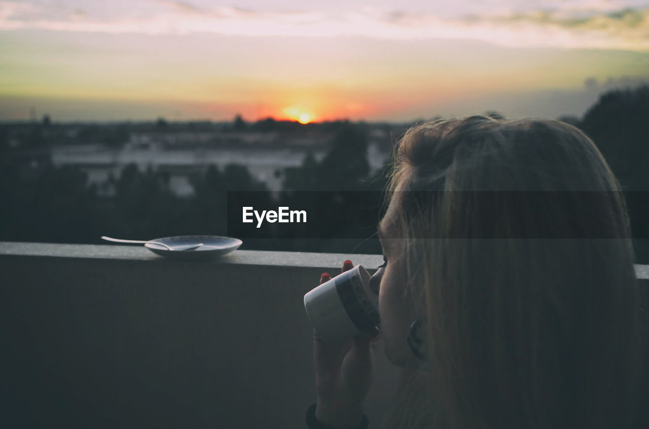 Teenage girl drinking coffee sitting at terrace during sunset