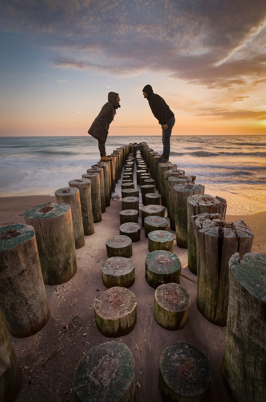 Side view full length of couple standing on wooden posts at beach during sunset
