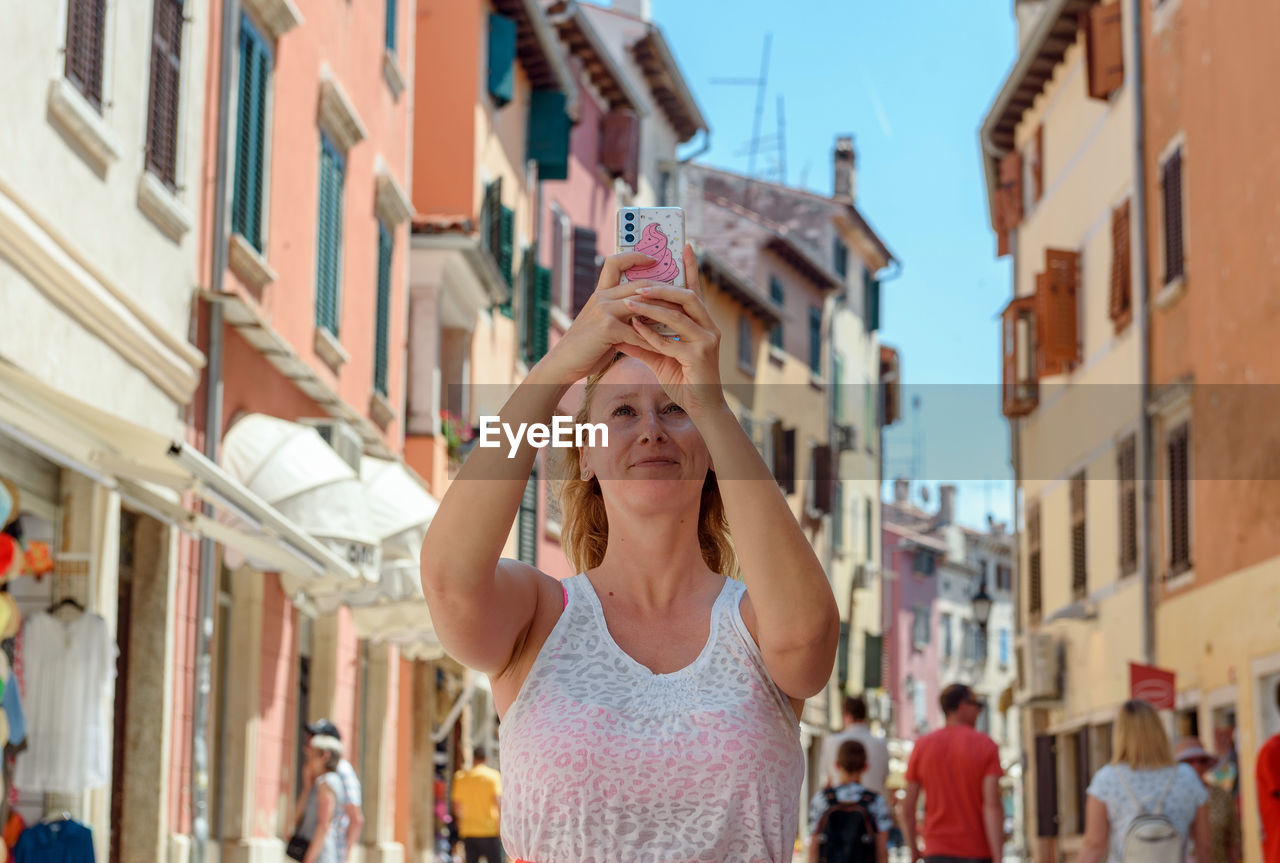 Front view of female tourist using mobile phone, taking photos in street of colorful town in rovinj