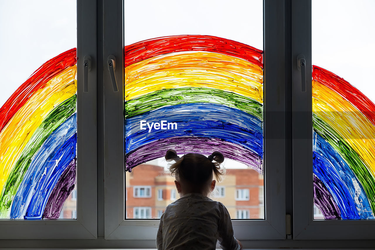 Little girl on background of painting rainbow on window. kids leisure at home. 