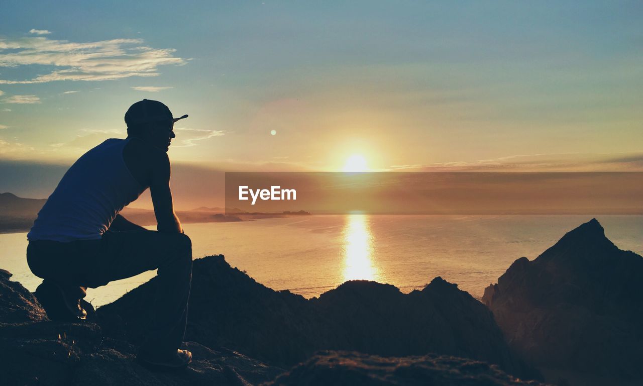 Rear view of man crouching on rock by seascape during sunset