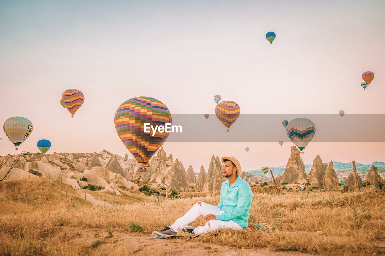 Thoughtful mid adult man sitting on field against hot air balloons flying in mid-air