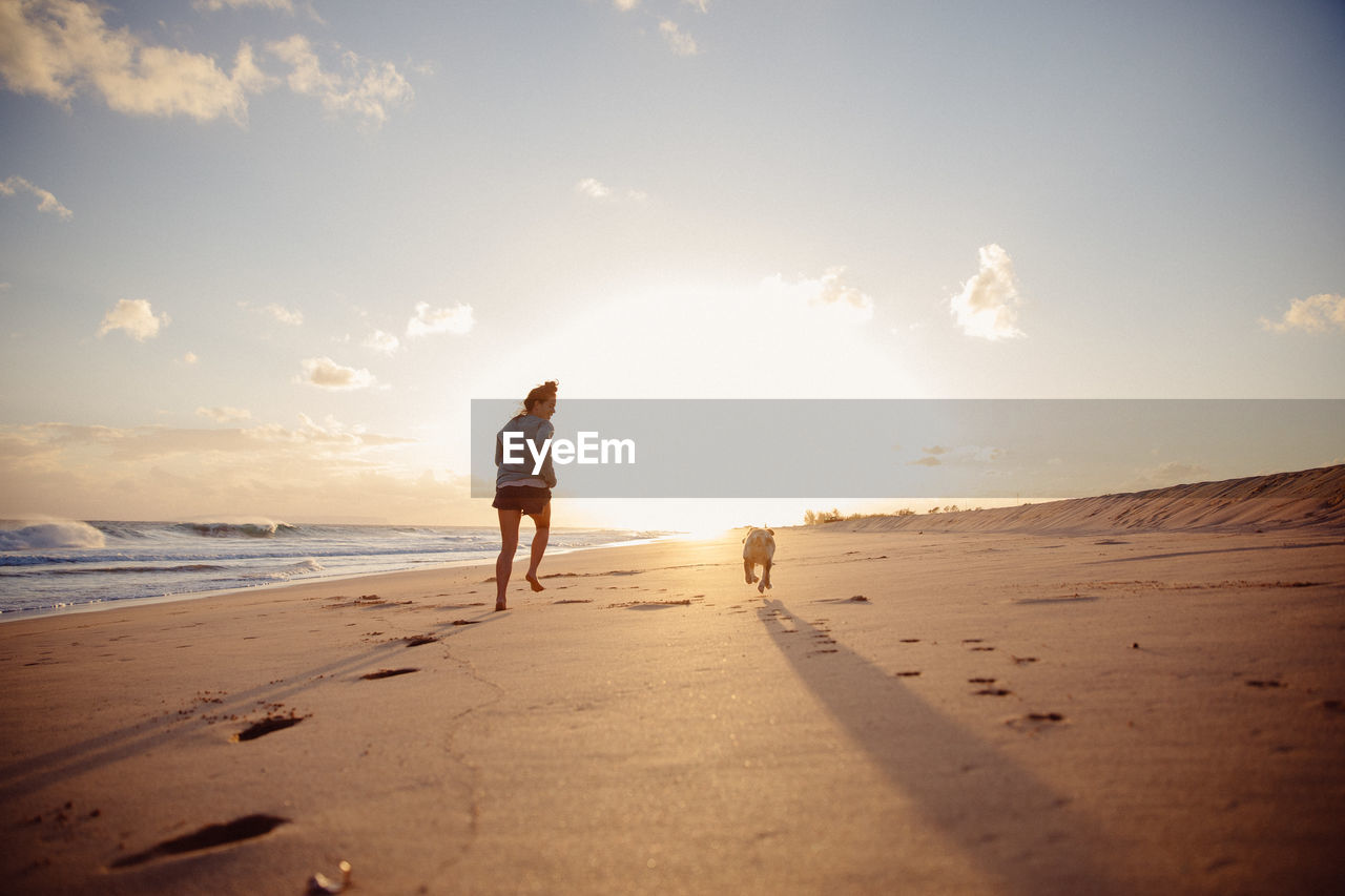 Rear view of woman with dog running at beach against sky