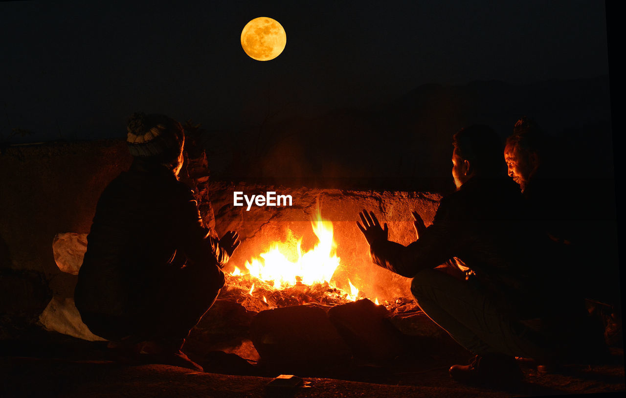 Rear view of men crouching by bonfire at night