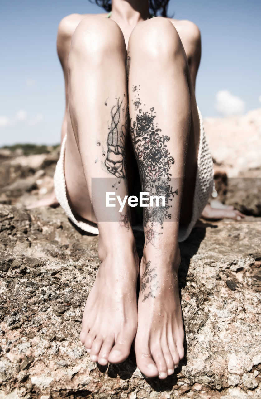 Low section of woman with tattoo sitting on rock against sky