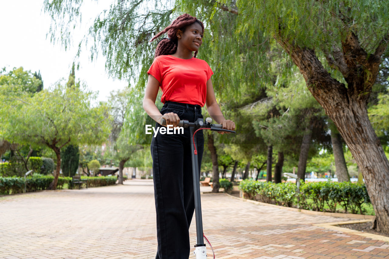 African american female riding electric scooter on paved walkway on street with green trees in city on summer day