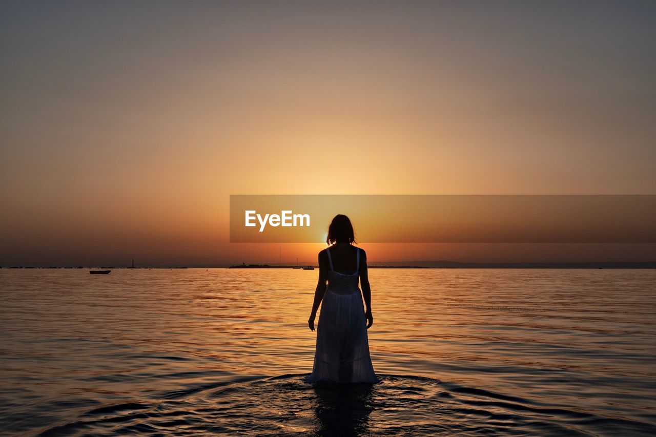 Rear view of woman standing in sea against sky during sunset