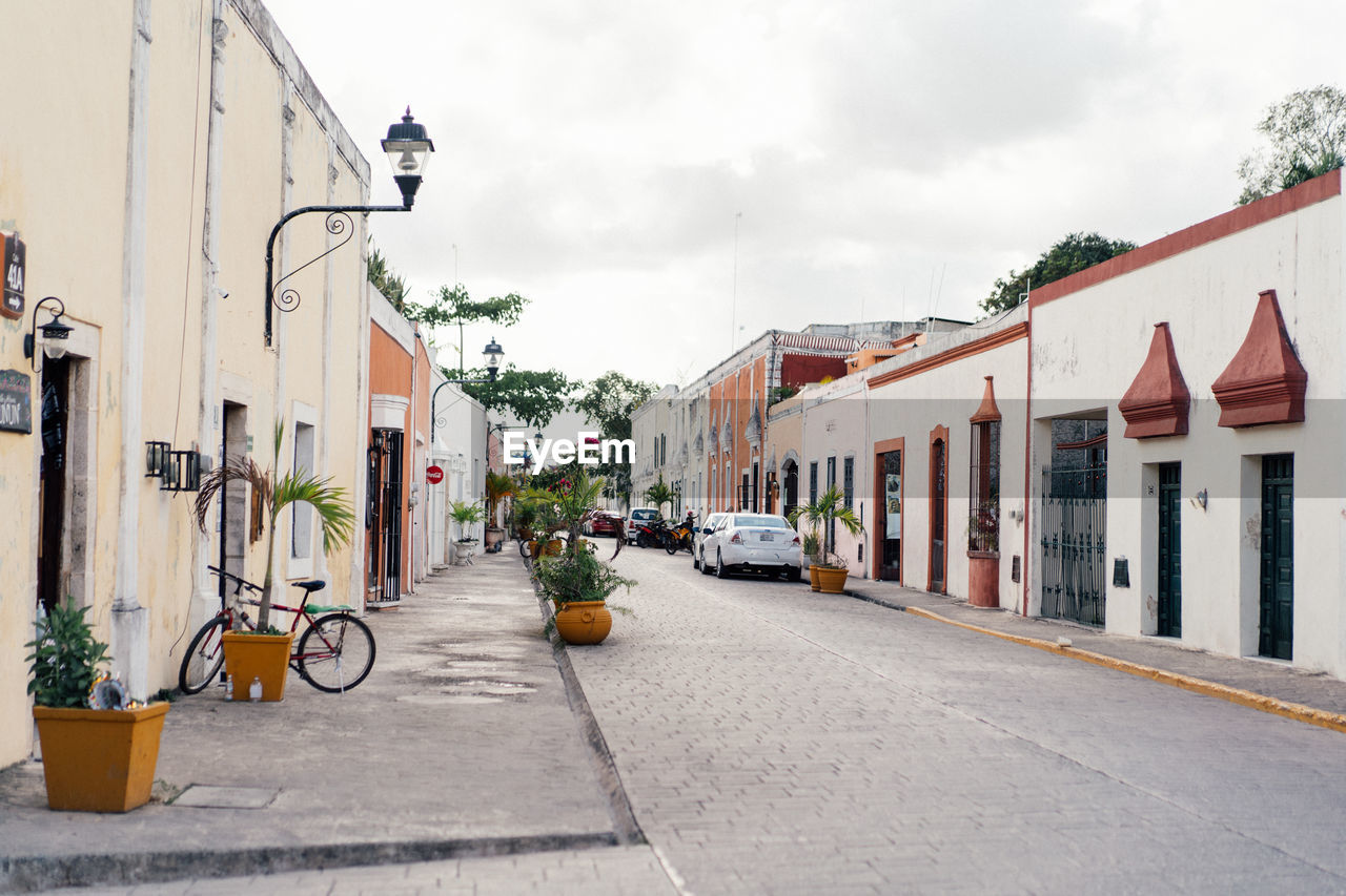 architecture, town, building exterior, neighbourhood, street, city, built structure, road, building, transportation, residential area, sky, nature, residential district, lane, cloud, day, mode of transportation, house, flowerpot, bicycle, infrastructure, potted plant, city street, no people, outdoors, plant, urban area, travel destinations, alley, houseplant, footpath, city life, vehicle, tree, car, motor vehicle