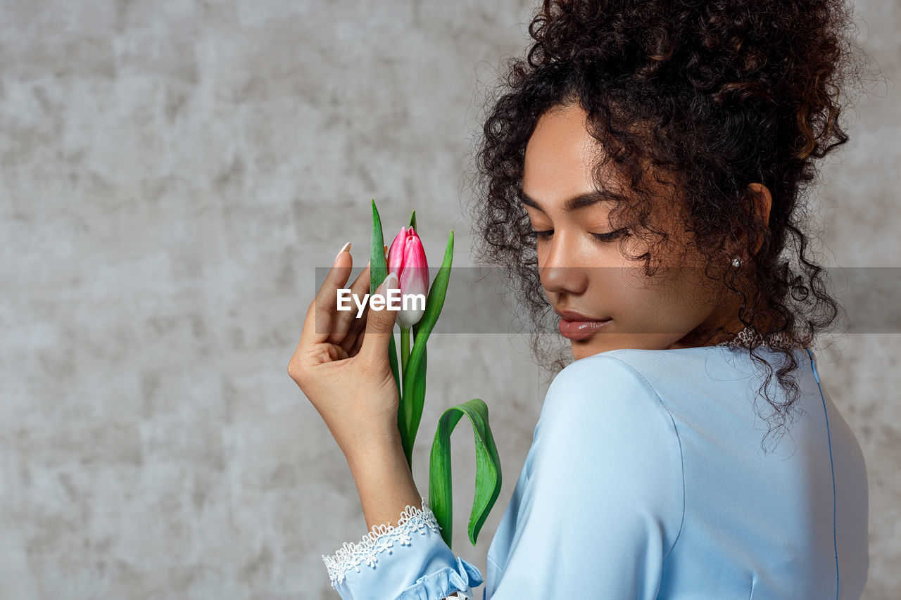 Young woman holding tulip while standing against wall