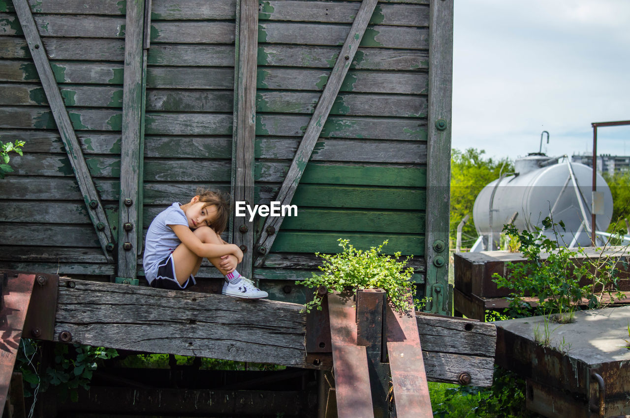 Sad girl sitting on abandoned wooden cargo container against sky