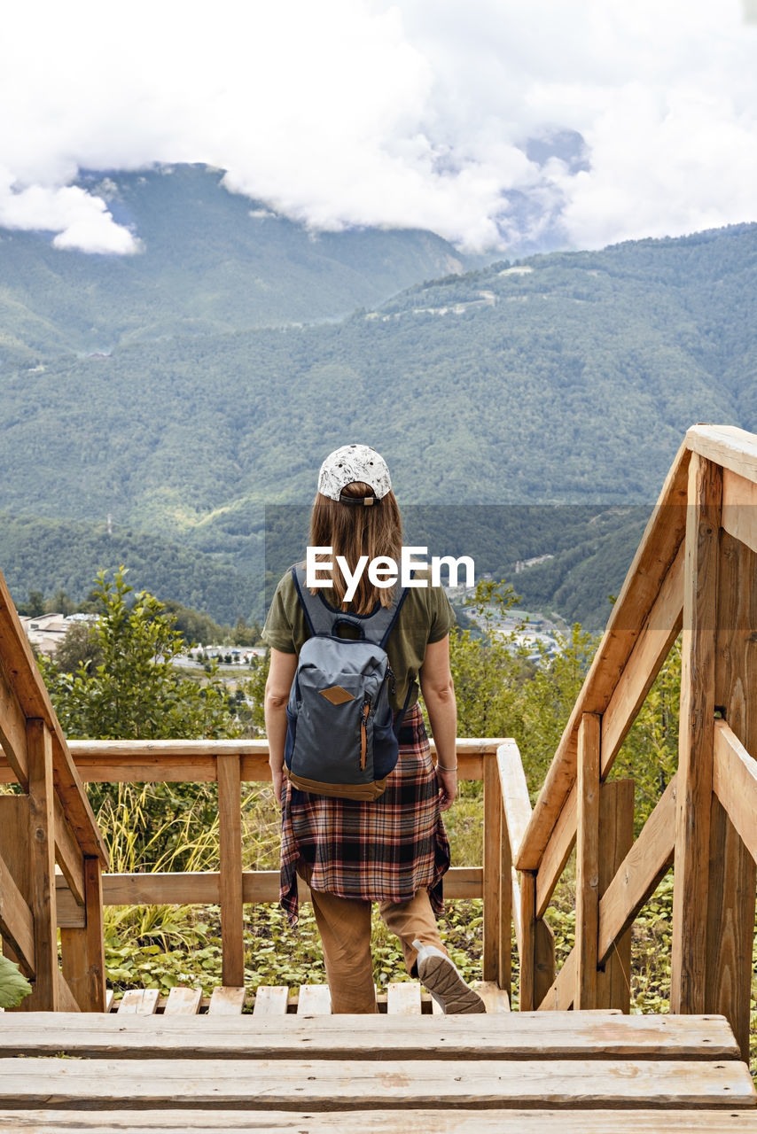 Rear view of young blonde woman looking at view of mountains standing on wooden viewpoint, hiking 