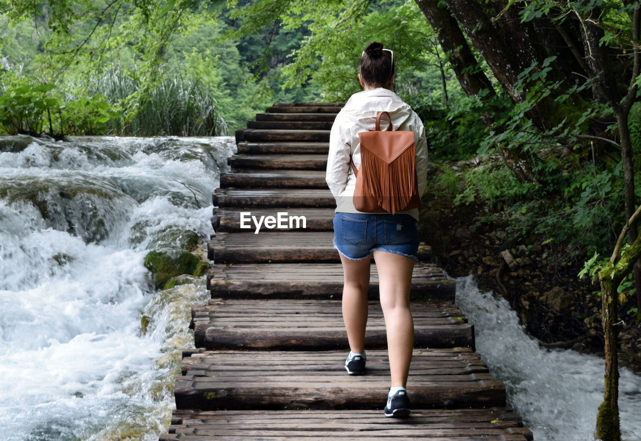 Rear view of woman walking on wooden overpass over river in forest