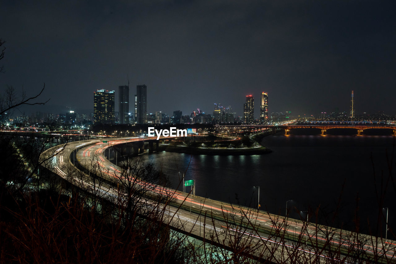 ILLUMINATED BRIDGE OVER RIVER AMIDST BUILDINGS IN CITY AGAINST SKY
