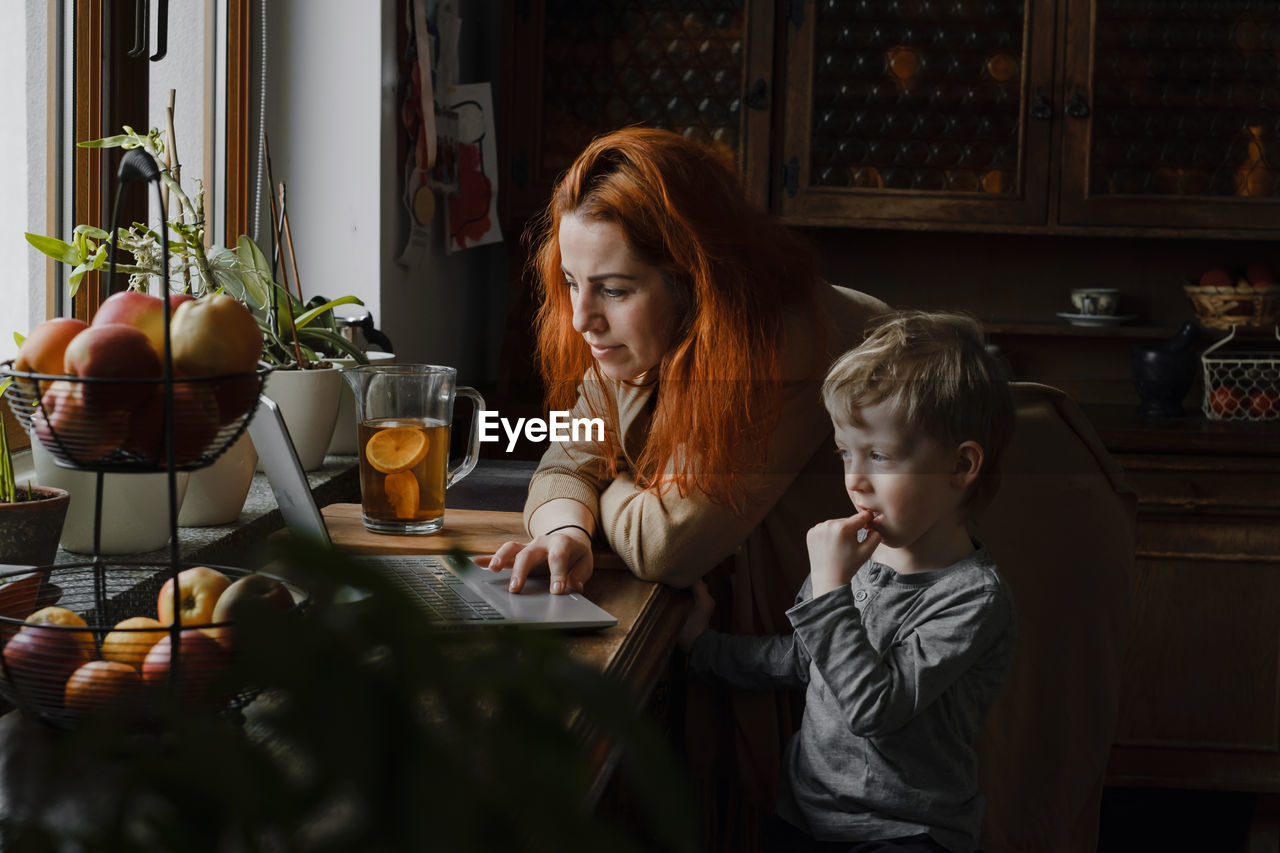 Ginger haired woman on kitchen with child looking in laptop. vintage house, cozy family life. boy