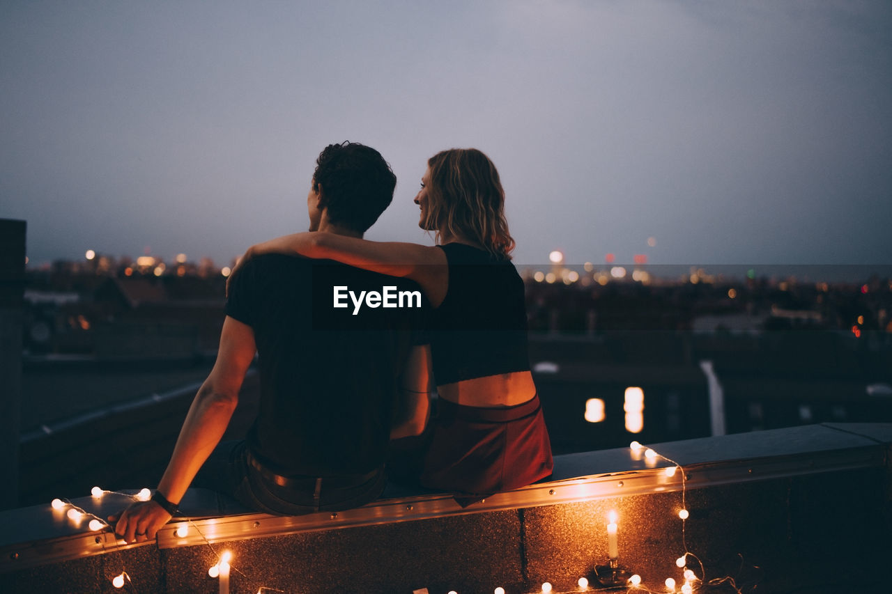 Rear view of couple with arm around sitting on illuminated terrace in city against sky