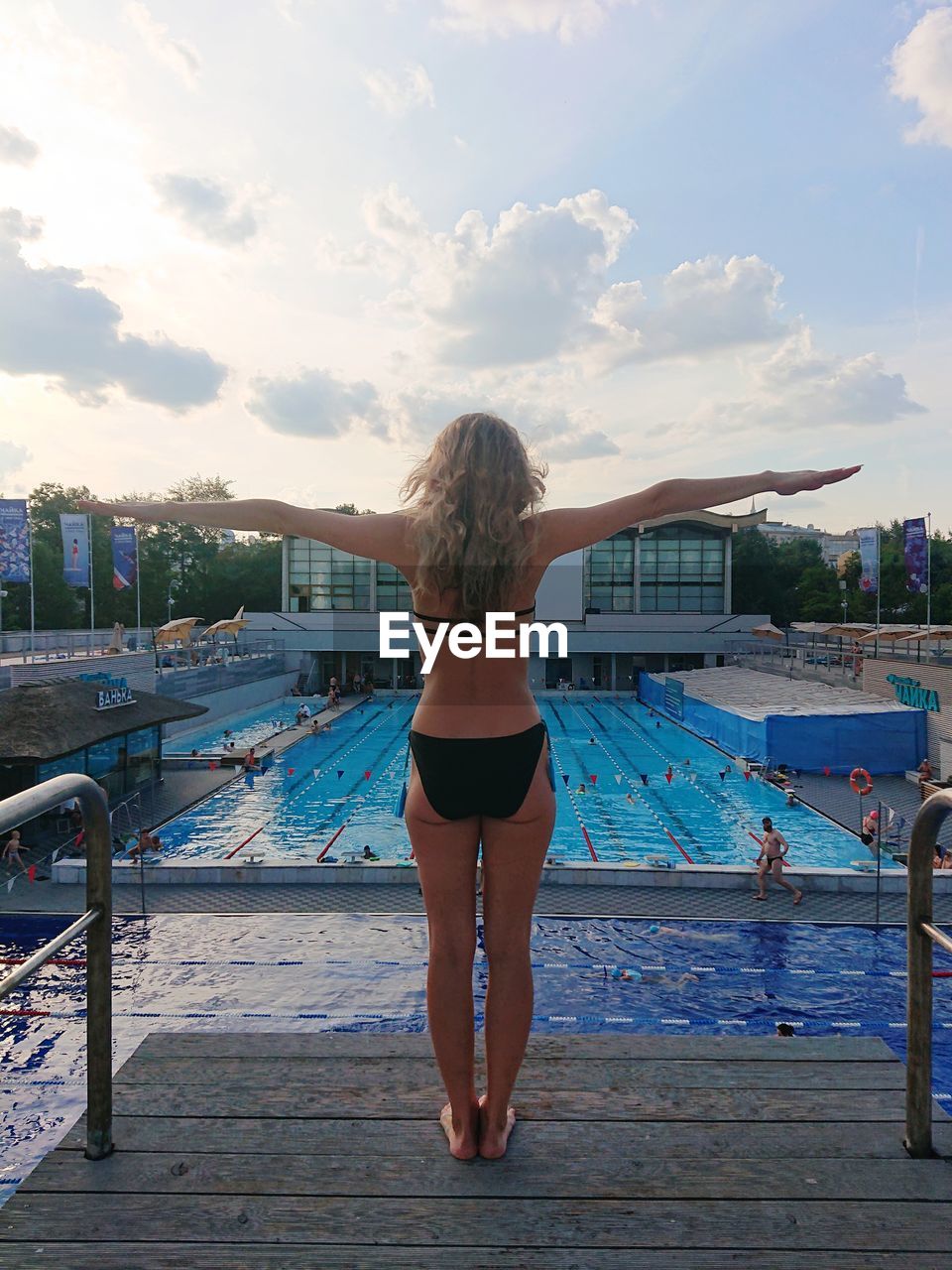 Rear view of woman with arms outstretched standing by swimming pool against sky