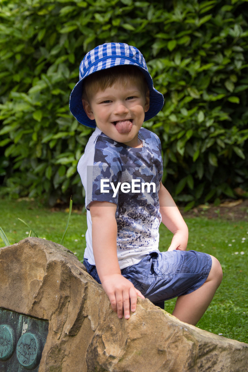 Portrait of boy sticking out tongue while sitting on rock at park