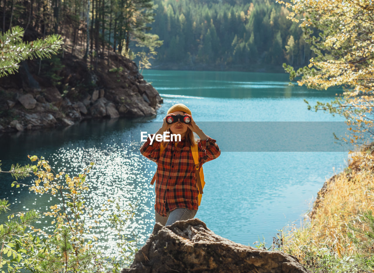 Young caucasian woman with binoculars by lake or river looking at birds migration in fall  birding