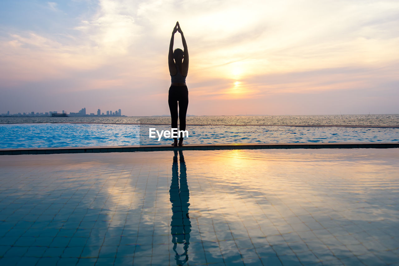 Woman exercising by sea against sky during sunset