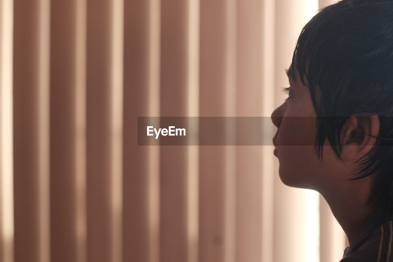 Thoughtful boy looking away while standing against curtain at home