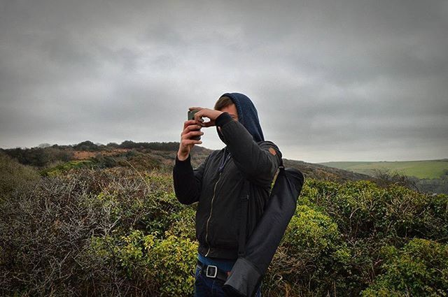 WOMAN PHOTOGRAPHING THROUGH SMART PHONE ON FIELD AGAINST CLOUDY SKY