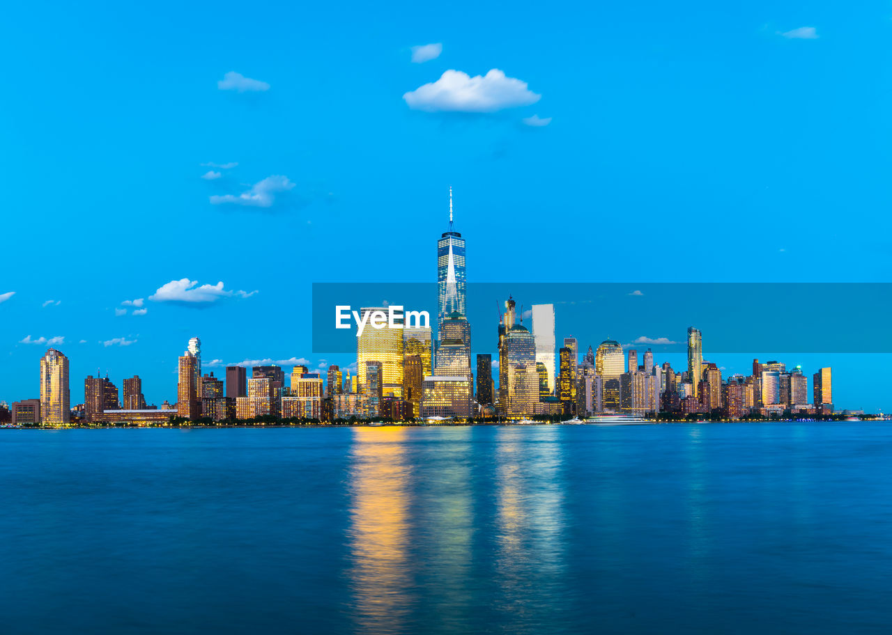 Illuminated one world trade center and buildings in city by hudson river against blue sky