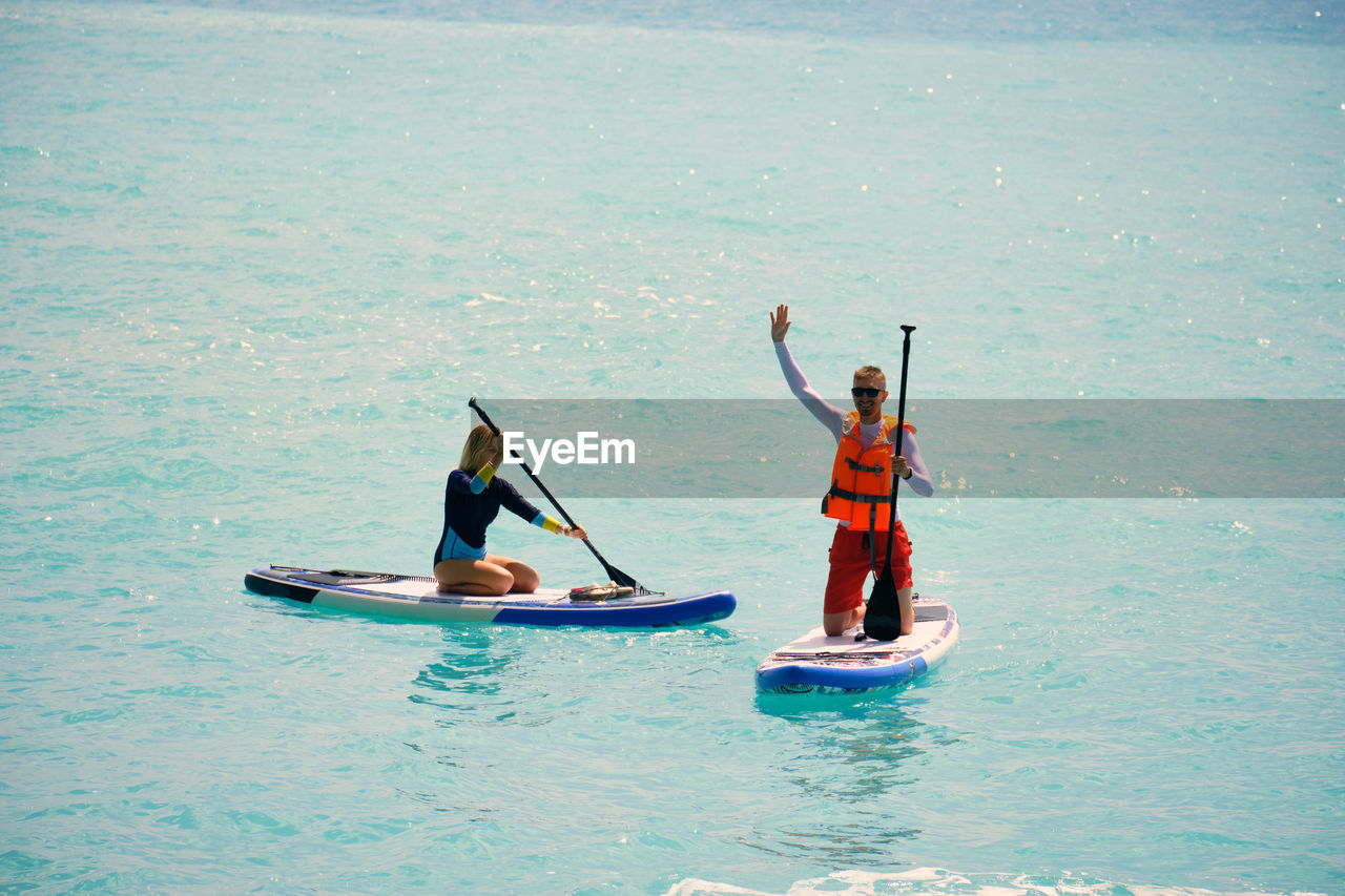 Young man dressed in swimsuit and lifevest waving and young woman paddling on stand up paddle board
