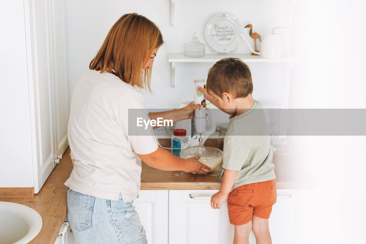Young woman mother and her toddler boy son cooking with blender at the table in kitchen at home