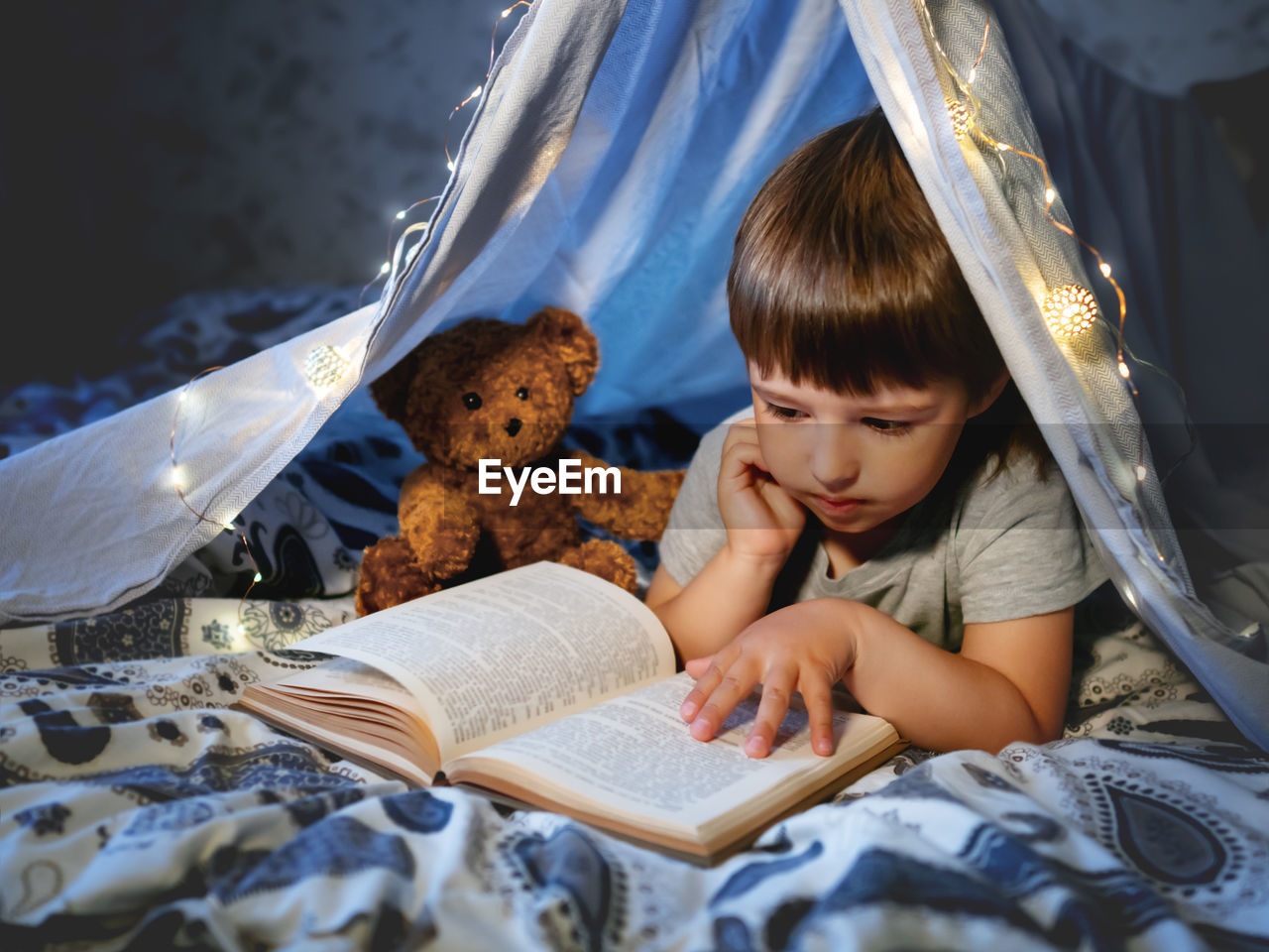 Little boy reads book. toddler plays in tent made of linen sheet with light bulbs on bed. 