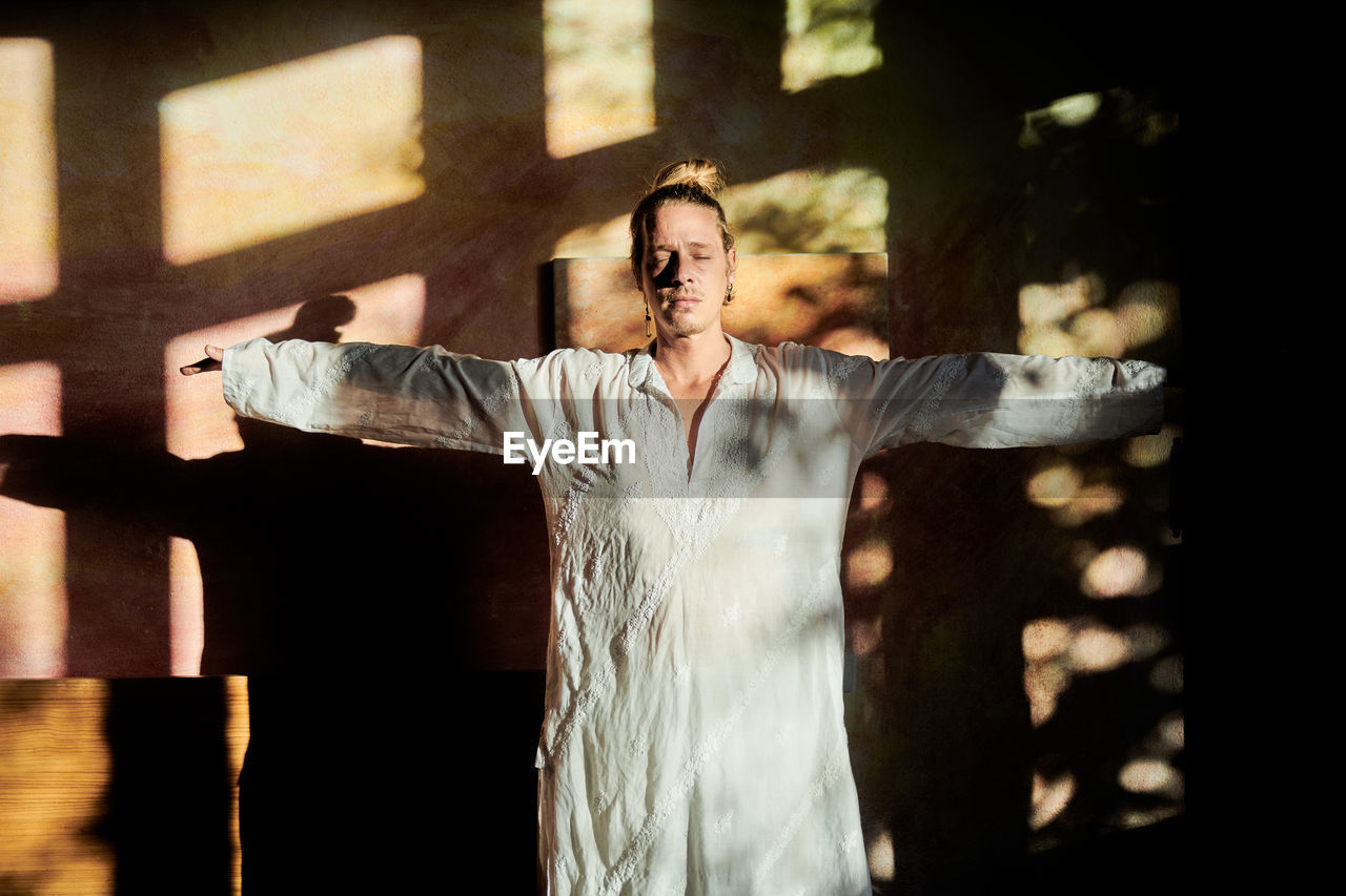 Young mindful male with stretched arms and closed eyes practicing yoga against wall with shades in soft sunlight