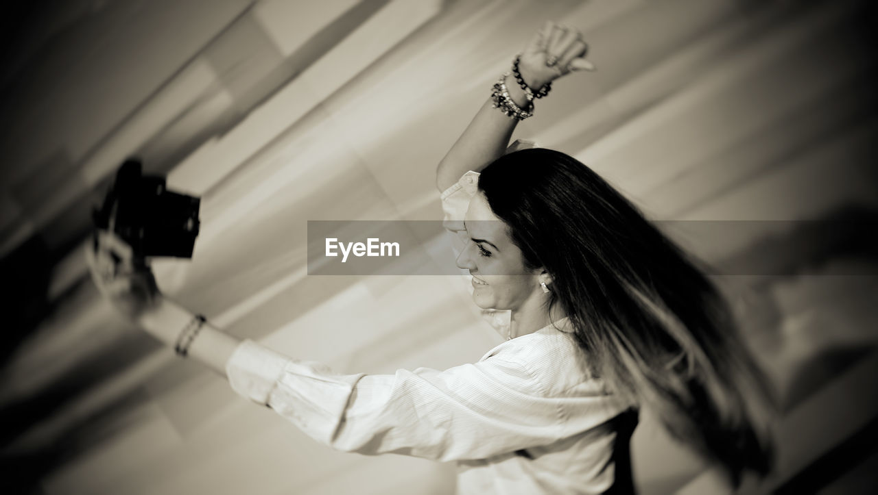 Side view of smiling young woman taking selfie while standing against wall