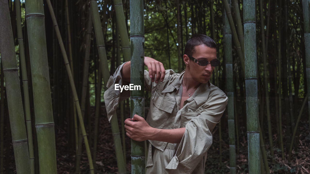 YOUNG MAN WEARING SUNGLASSES STANDING BY TREE TRUNKS