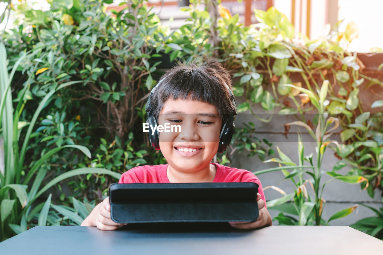 Asian cute boy wearing wireless headphones and smiling happily while playing games on the tablet.