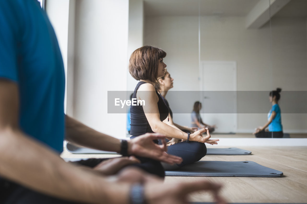 Mirror reflection of group of people with female instructor practicing yoga meditation in lotus pose with mudra hands during class in studio