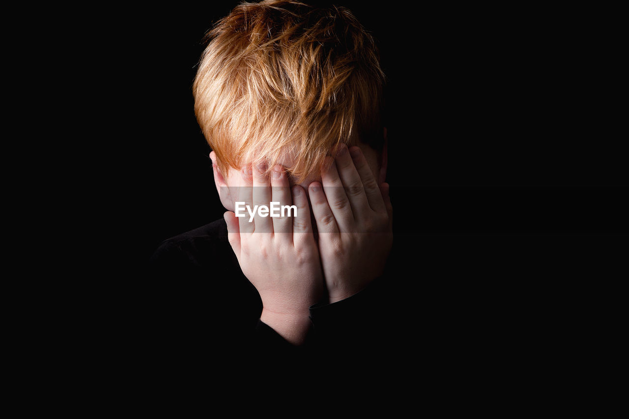 Close-up of boy covering face over black background
