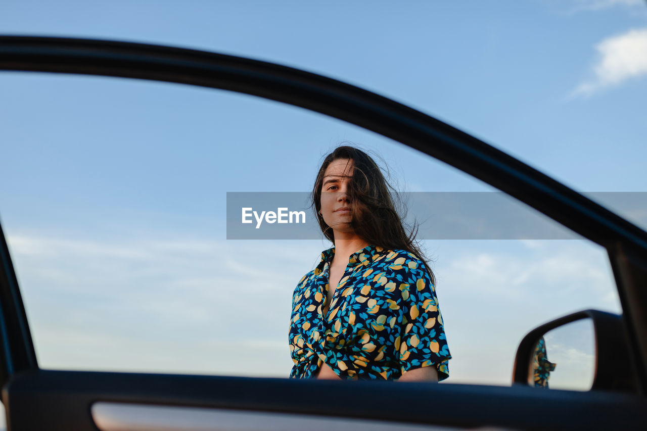 From below of young charming female in trendy wear with ornament looking at camera through glass auto door under blue cloudy sky