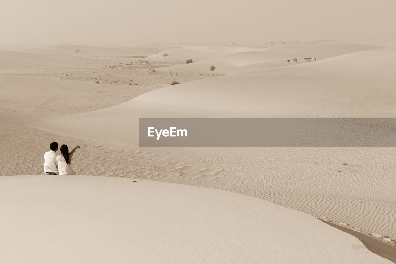 Rear view of couple sitting on sand at desert during sunny day