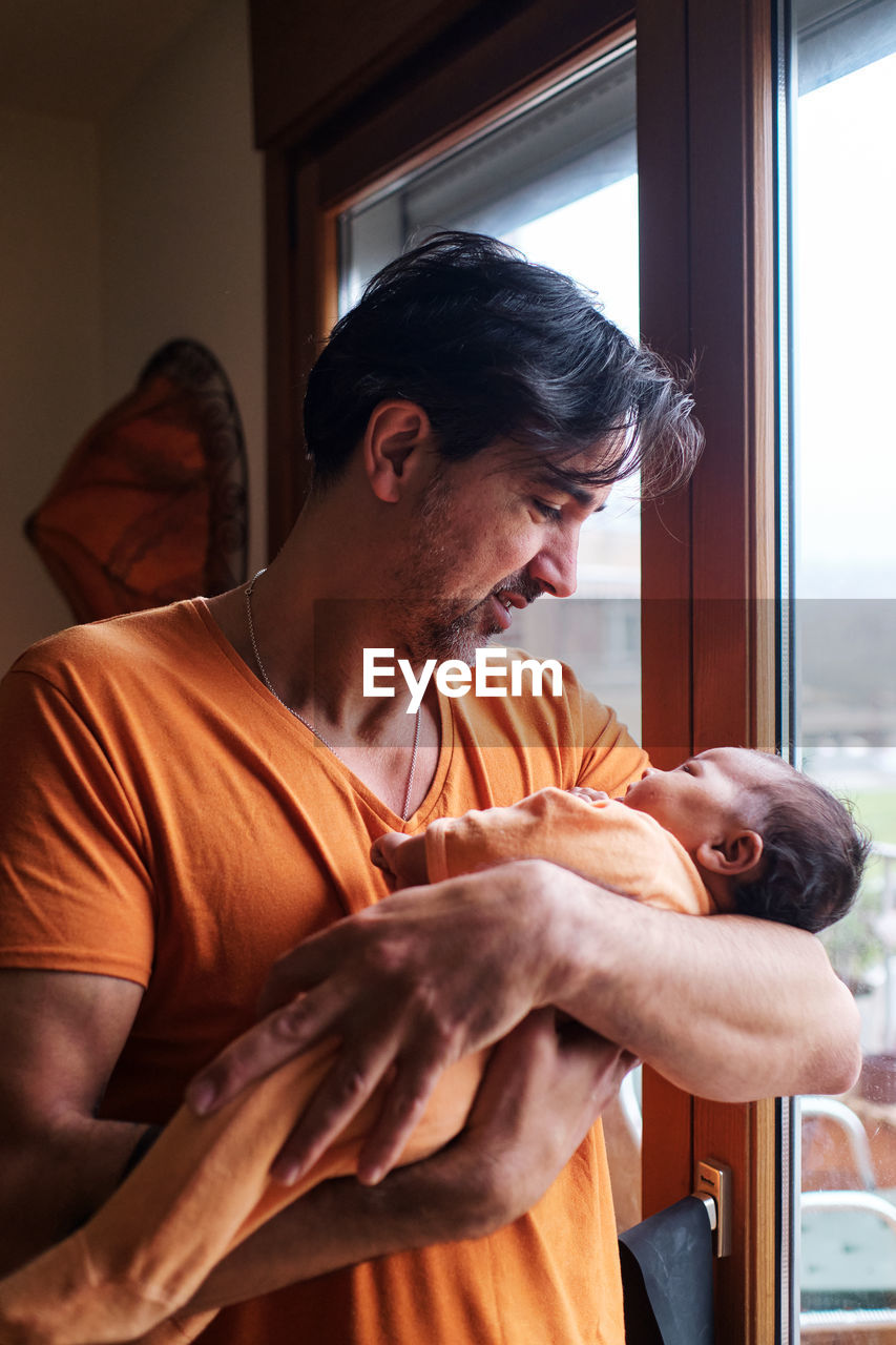 Bearded mature hispanic dad gently holding cute little baby while standing near glass door at home