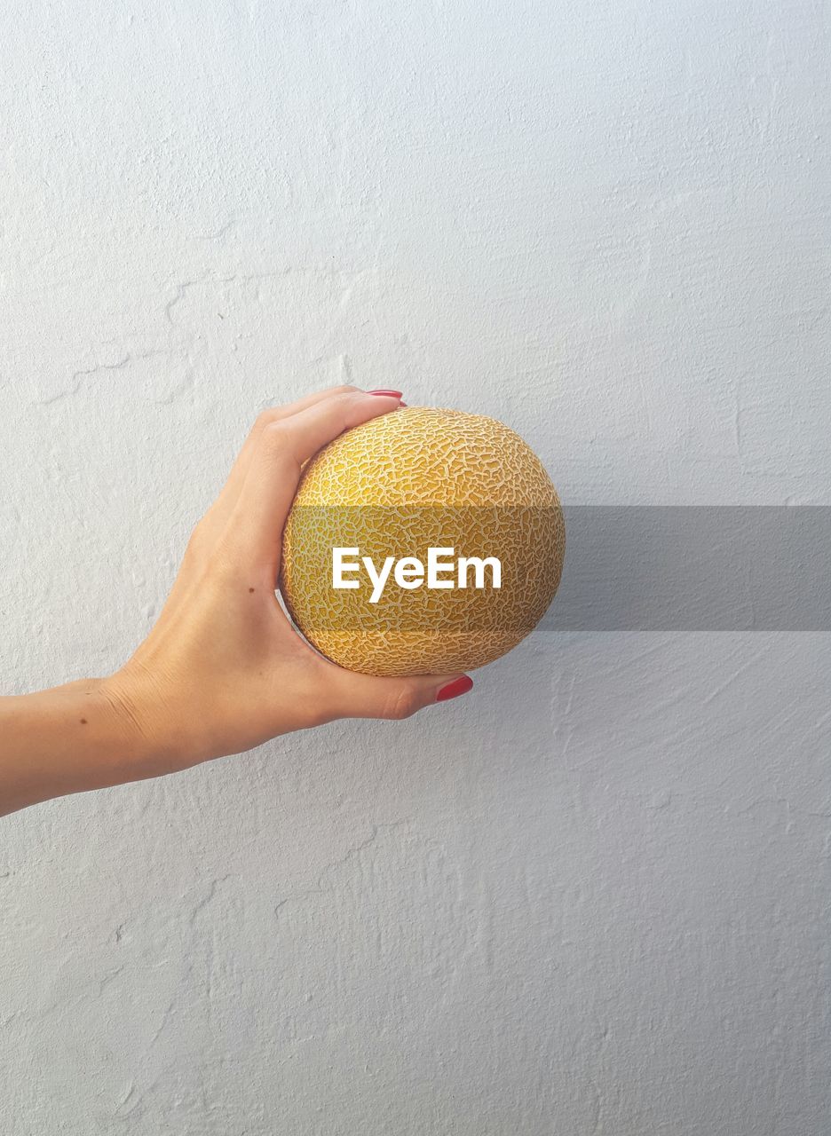 Cropped hand holding muskmelon against white wall